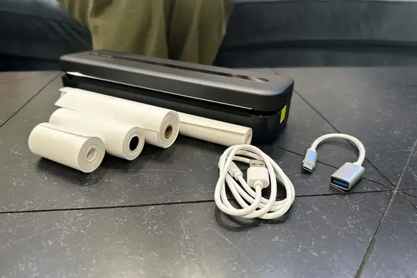 A portable printer on a table with several paper rolls a USB cable and an adapter