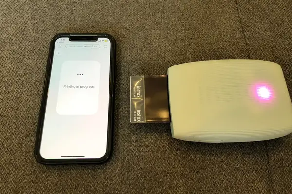 A smartphone displaying Printing in progress next to a Fujifilm Instax photo printer outputting an image