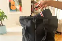 A hand placing a small bag with a striped strap into a larger black tote bag with a house interior in the background