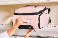 A person putting the Calpak Luka Soft-sided Mini Carry-on Luggage in an overhead compartment of a plane