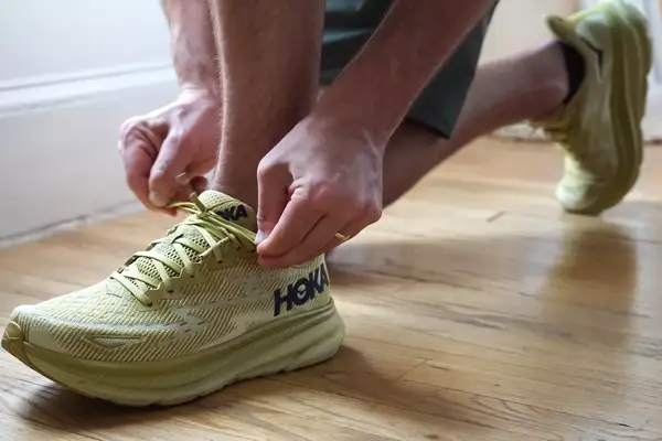 A person kneeling on the ground while tying the shoelace of a Hoka Clifton 9