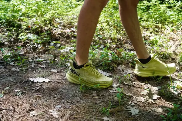A close-up of two legs wearing Hoka Clifton 9 sneakers while walking in dirt