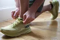 A person kneeling on the ground while tying the shoelace of a Hoka Clifton 9