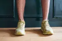 A close-up of two feet facing the camera while wearing Hoka Clifton 9 sneakers indoors on a wooden floor