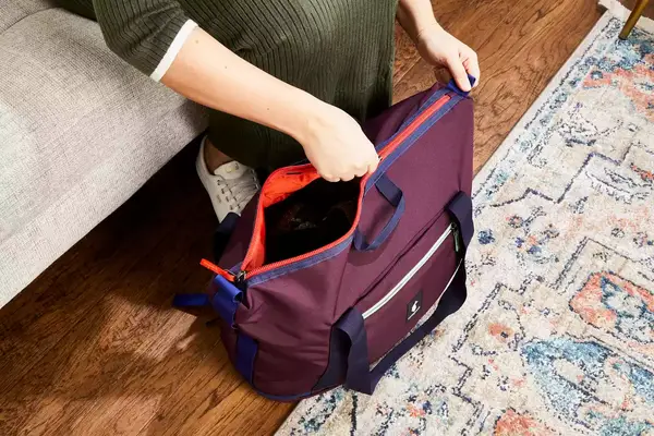 Hands zipping the Cotopaxi Viaje 35L Weekender Bag - Cada Día on the floor