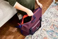 Hands zipping the Cotopaxi Viaje 35L Weekender Bag - Cada Día on the floor