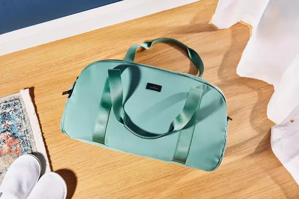 The Arlo Skye The Weekender bag on a wooden floor next to shoes and a rug