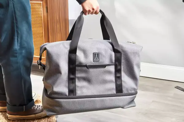A hand holding the handles on the Travelpro x Travel & Lanetrek Drop-bottom Weekender Bag