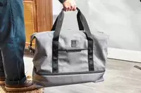 A hand holding the handles on the Travelpro x Travel & Lanetrek Drop-bottom Weekender Bag