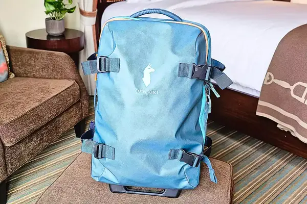 A blue Cotopaxi Allpa 65L Roller Bag with straps and a handle placed on a bench in a hotel room setting