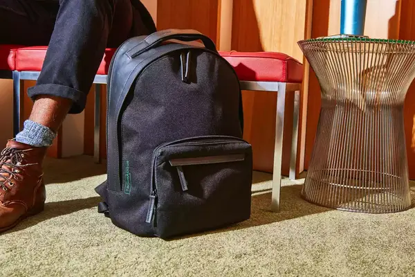 The Troubadour Orbis 1-Pocket Backpack on the floor in front of a bench with a leg and foot in the background