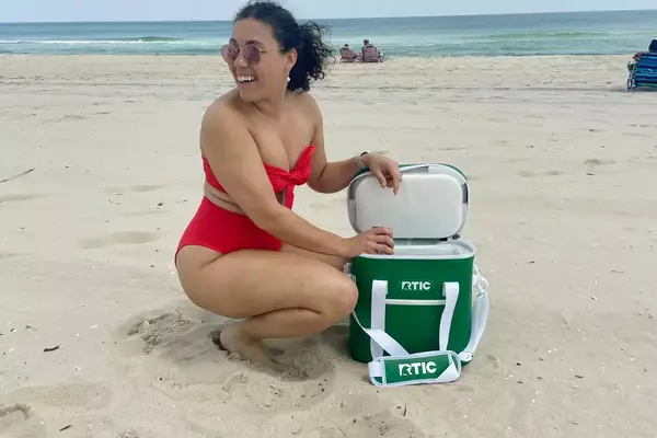 A person on a beach removes a canned beverage from the RTIC Insulated Soft Cooler Bag