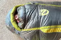 A person peeks out of the Nemo Coda Genderless Endless Promise Down Mummy Sleeping Bag