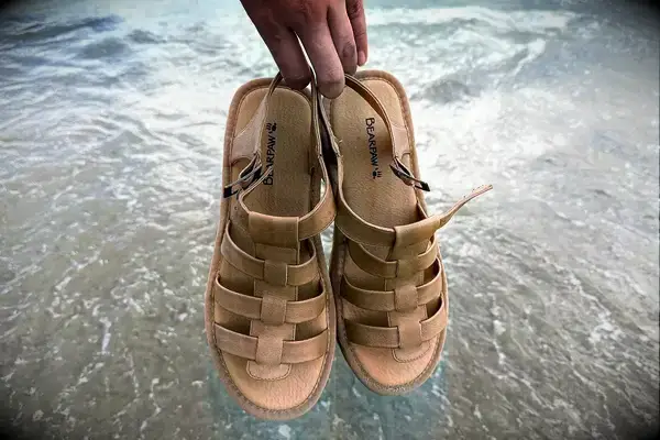 A person holding Bear Paw High Seas Iced Coffee Sandals in front of water