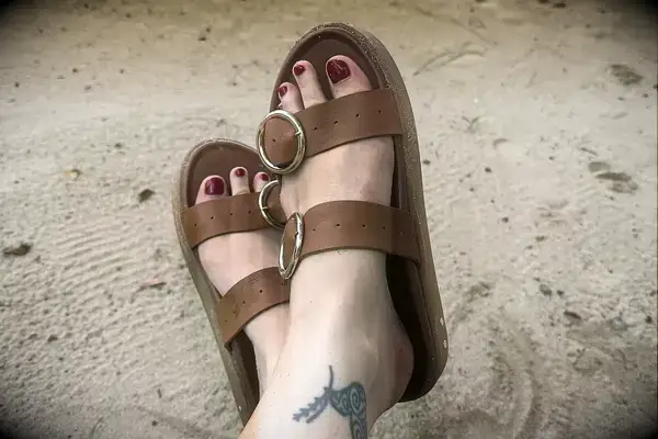 A person wearing the Beek Warbler Leather Platform Platform Leather Slides with their feet crossed and a sand background