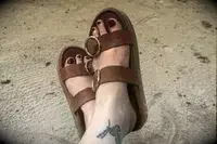 A person wearing the Beek Warbler Leather Platform Platform Leather Slides with their feet crossed and a sand background