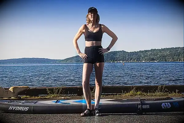 Person holding a paddle and standing near a green kayak by a lake wearing the Sheila Nellie Scoop Bikini