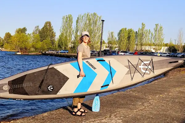 A person holding the Hydrus Paradise Touring Inflatable Paddle Board while standing outside of the water