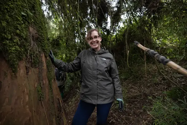 A person standing in a forested area, leaning against a tree trunk with a smile, wearing outdoor attire