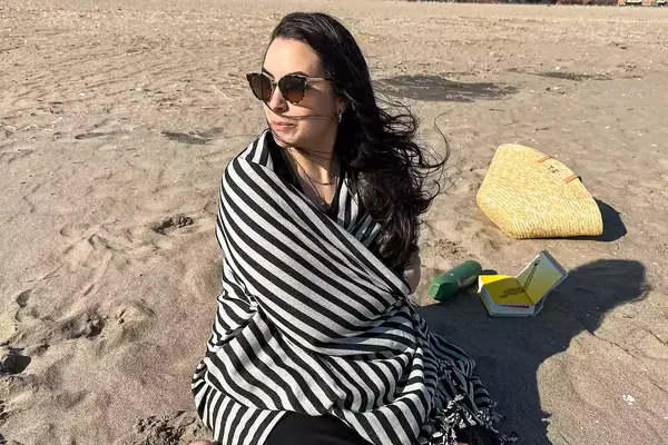 A person sitting on a beach wrapped up with the Fairmont Turkish Linen Towel
