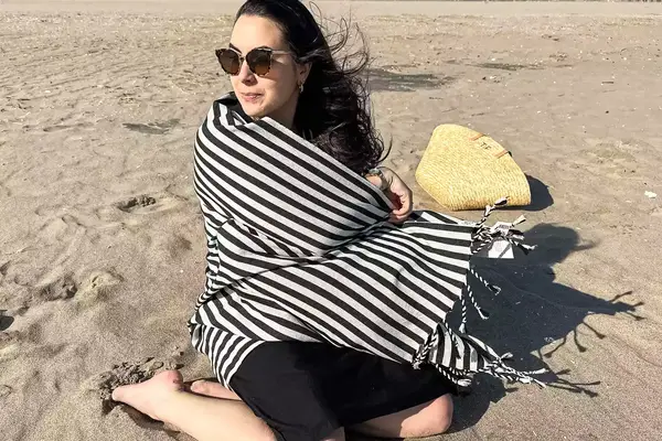 A person sitting on a beach wrapped up in the Fairmont Turkish Linen Towel