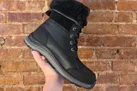 A hand holds up an UGG Adirondack III Boot in front of a brick wall