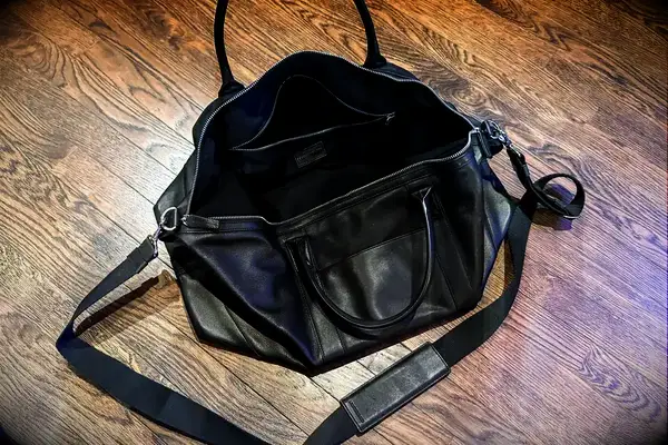 An open leather bag with handles and a shoulder strap placed on a wood floor