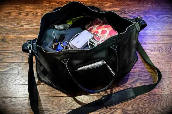 An open black bag on a wooden floor holding keys a mobile charger wires and other assorted items