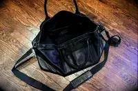 An open leather bag with handles and a shoulder strap placed on a wood floor