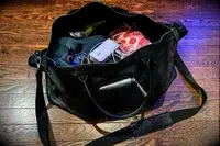 An open black bag on a wooden floor holding keys a mobile charger wires and other assorted items