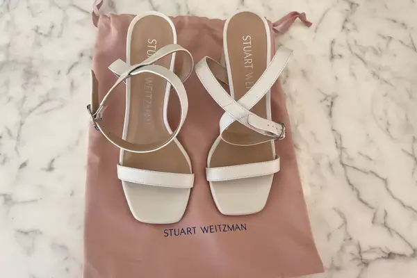 The Stuart Weitzman Nearlynude Strap Sandals on top of the shoe bag on a white marble countertop