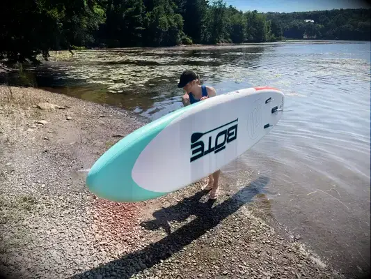 A person standing on the SereneLife Inflatable Stand-up Paddleboard while in a lake