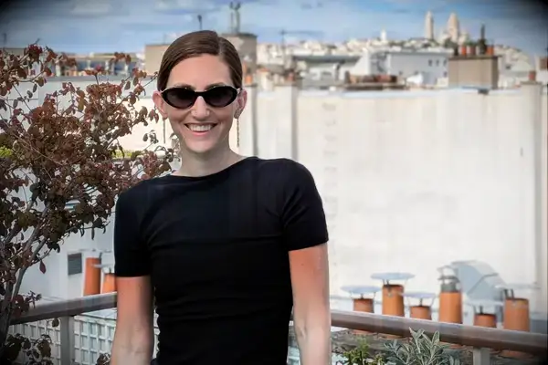 A woman smiling and wearing sunglasses, standing on a balcony with a city view in the background