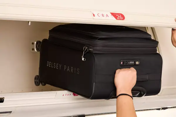 A person places the Delsey Paris Cruise 3.0 Soft Carry-On Expandable Spinner into an overhead compartment