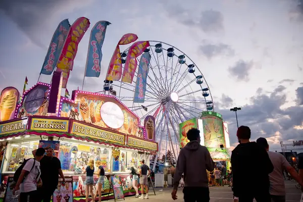 People walking near food stands and a Ferris wheel at a fair