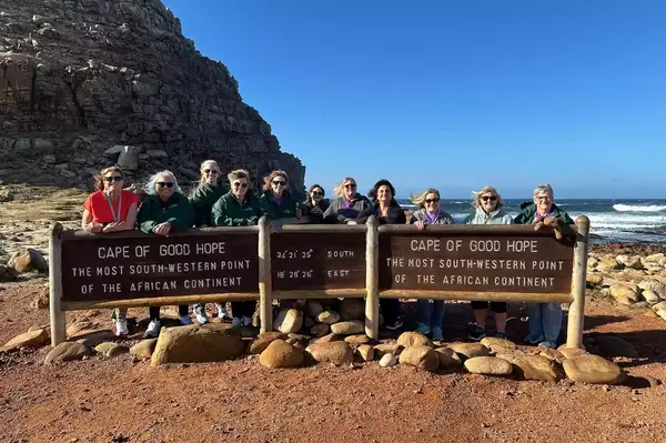 A group of women gathered at the sign for Cape of Good Hope, the southernmost point of Africa, standing on a rocky terrain with the ocean visible