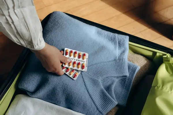 A persons hand placing a blister pack of pills onto a folded sweater in an open suitcase