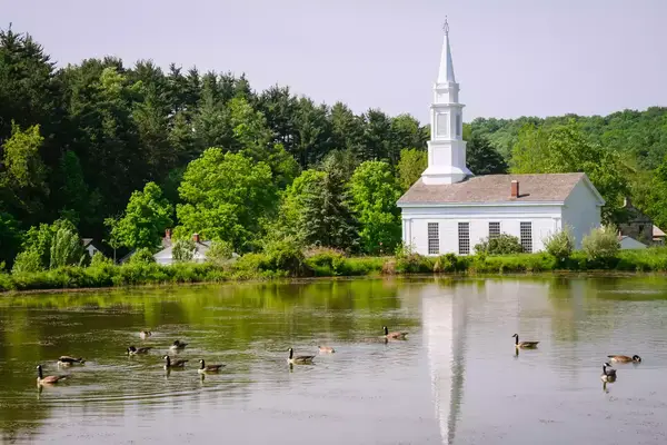 Church with a steeple by a pond, surrounded by trees, with geese swimming in the water.