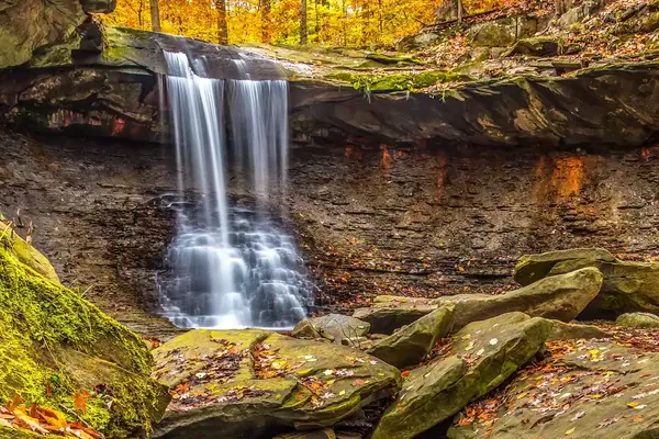 Waterfall flowing over a rocky ledge in a forest setting, surrounded by rocks and autumn foliage.