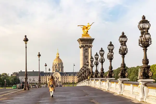 Person walking on a bridge in Paris with ornate lamp posts and domed building in the distance