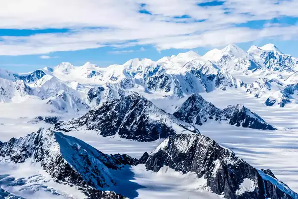 Snow-covered mountain peaks with glaciers in a rugged landscape, partly cloudy sky above