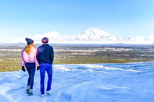Two people standing on a snowy landscape overlooking a forest and distant mountains in clear weather