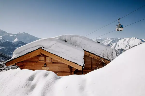 A snow covered, wooden chalet sits in the sunshine with a ski lift in the background, with a bright blue ski and snow covered Swiss mountains.