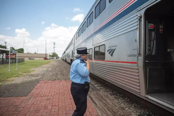 A train conductor in a blue shirt and navy hat stands on a brick platform looking in towards a bi-level Amtrak train car.