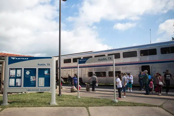 A bi-level silver Amtrak train sits in the station as passengers stand outside, with Austin, Texas, signage in the foreground.