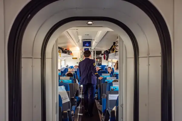 Interior view of a train carriage with passengers seated and a crew member standing in the aisle, as seen through a doorway
