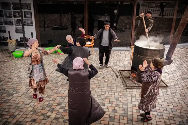 Group of people dancing and others cooking outdoors with a large pot on a fire