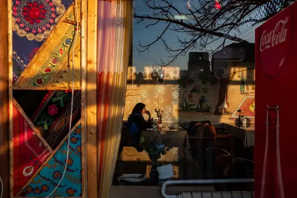A person sitting inside a cafe as seen through the window, surrounded by Uzbek decorations and reflections of the surroundings on the glass