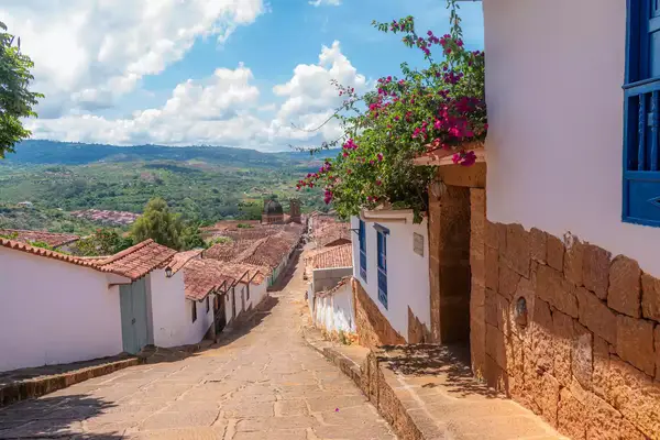 A cobblestone street descends among white buildings adorned with flowers, opening to a view of a scenic countryside landscape