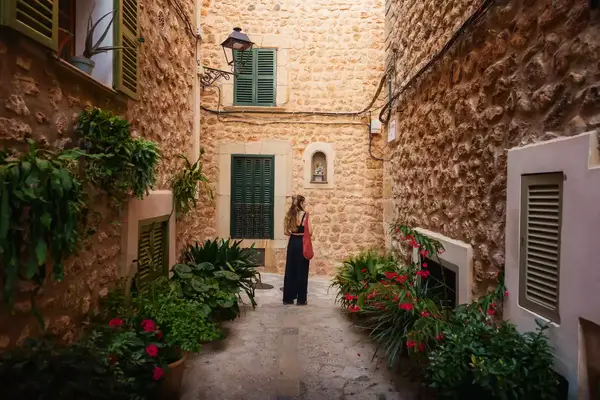 A woman in a narrow stone alleyway surrounded by plants, back facing the camera